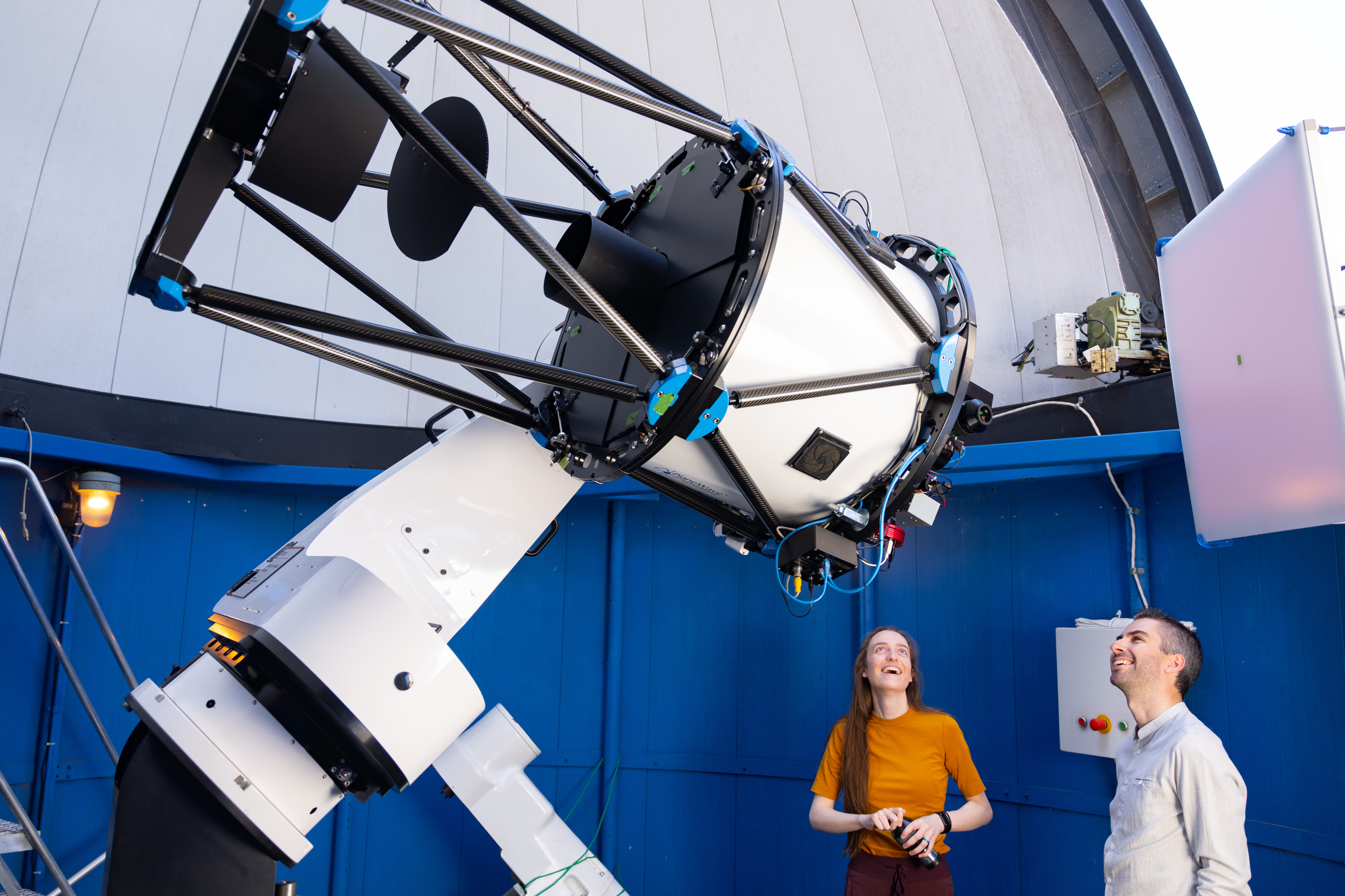 Image inside the Burke-Gaffney Observatory dome with white ceilings and blue coloured walls. The white and black telescope is being pointed to by Tiffany Fields, Astronomy Technician, wearing yellow and Dr. Vincent Henault-Brunet looks onward as well, wearing white.